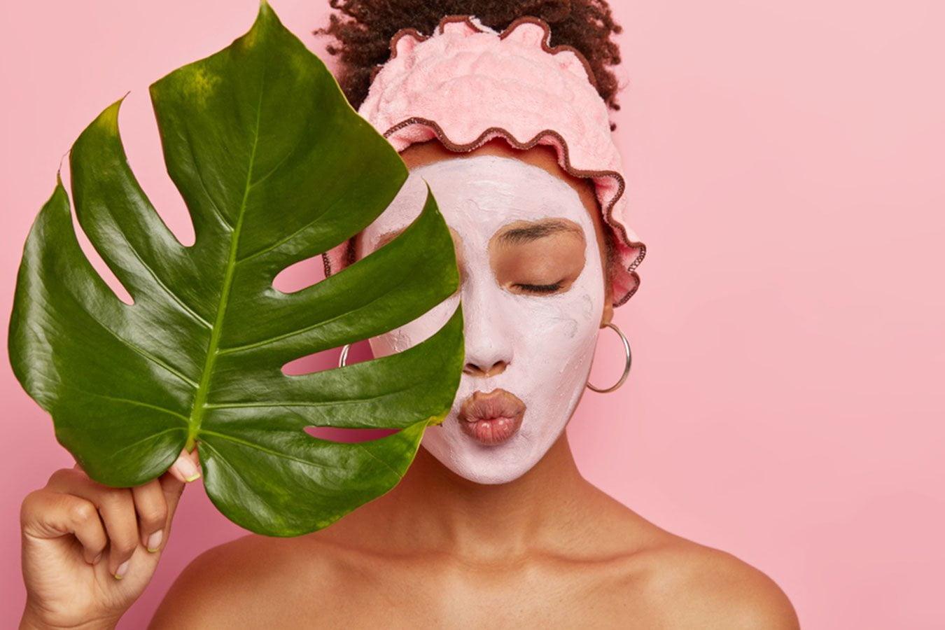 Woman with clay face mask, pink headband, holding green leaf against pink background
