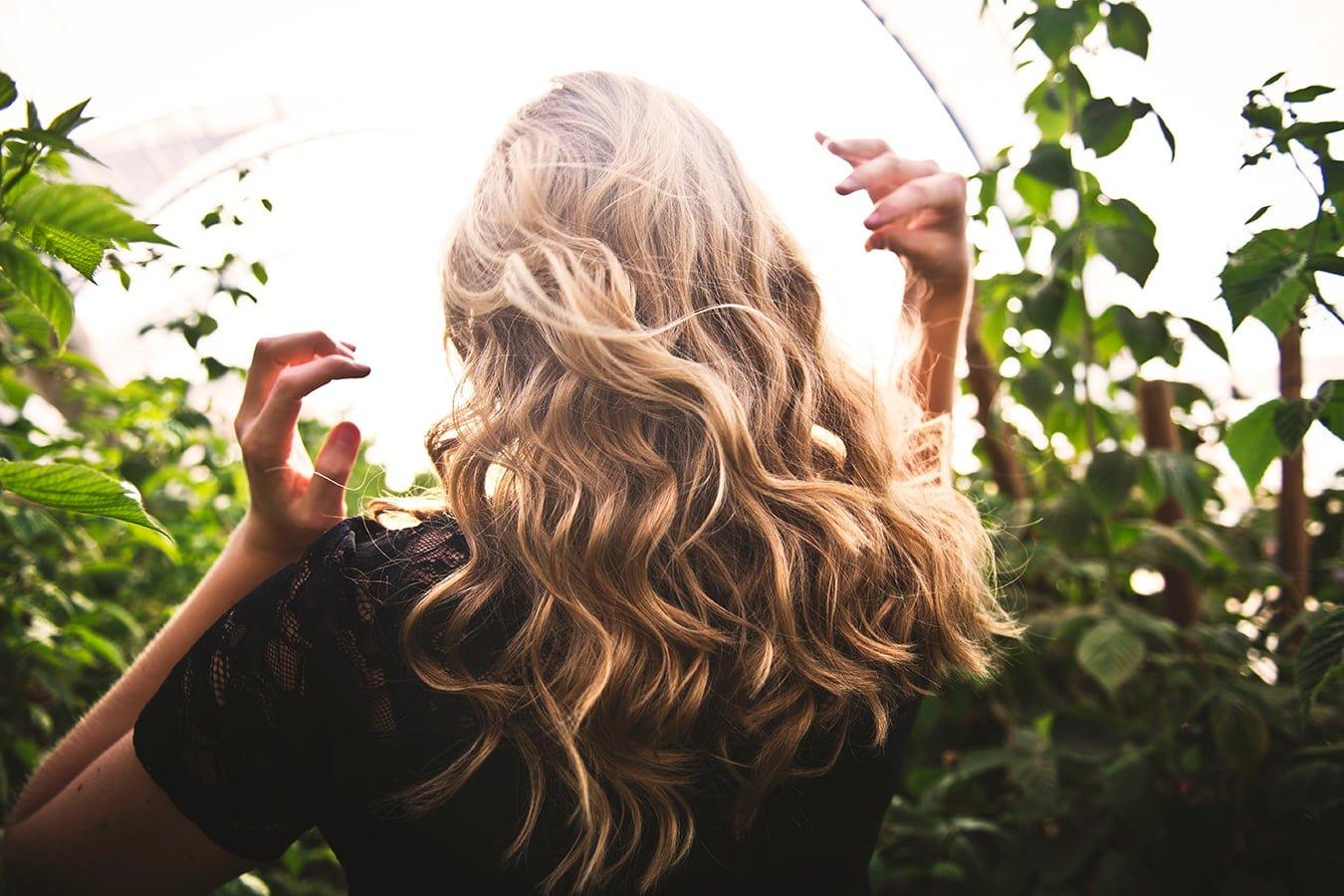 Woman with healthy wavy blonde hair outdoors surrounded by green plants, haircare concept