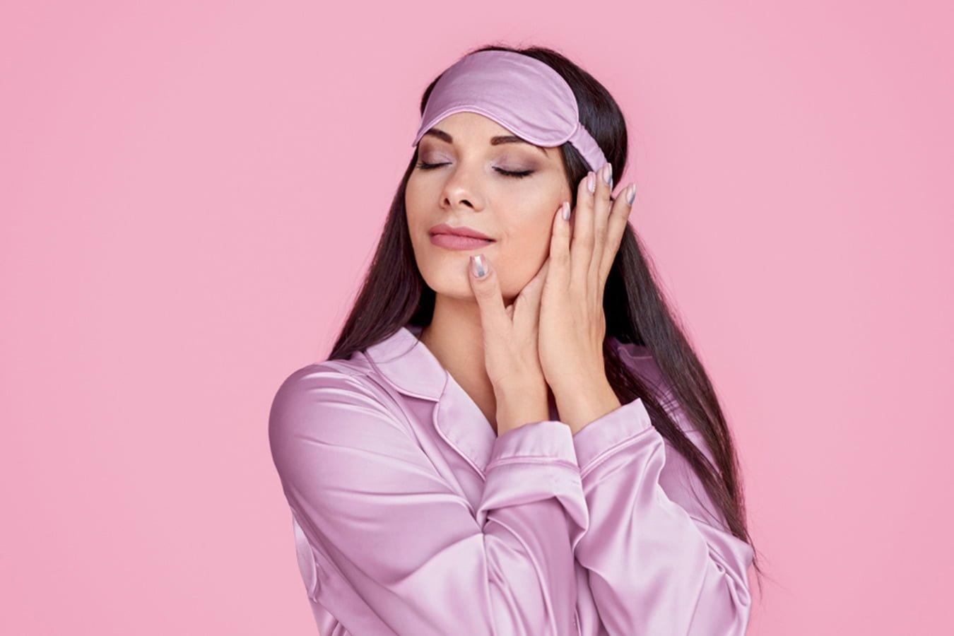 Woman in pink satin sleepwear and eye mask posing against pink background