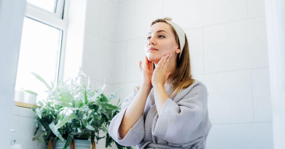 Woman applying skincare in bright bathroom, wearing headband and robe, with plants nearby