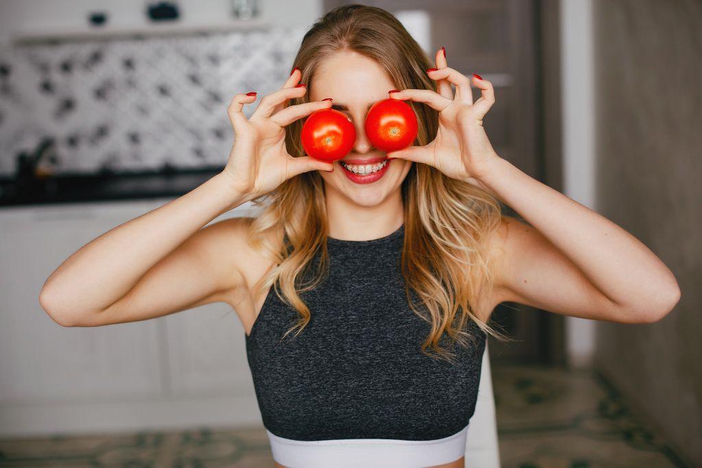 Smiling woman holding fresh tomatoes over eyes in kitchen, natural skincare concept