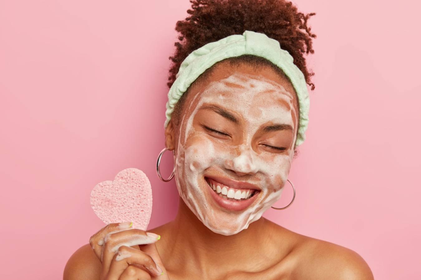 Smiling woman with foaming face cleanser, wearing headband, holding pink heart sponge
