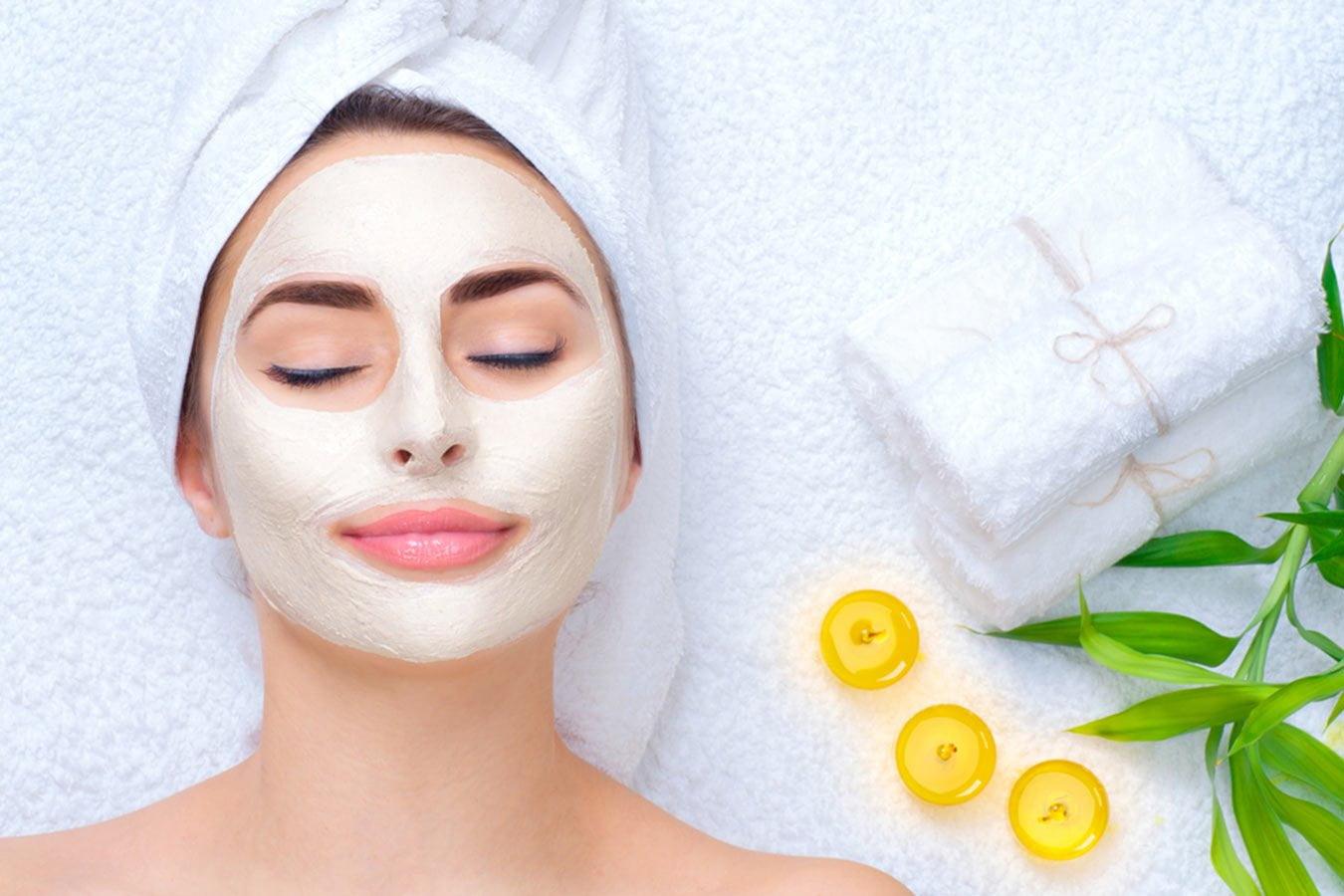 Woman relaxing with clay face mask, wrapped in towel, spa towels, candles, and green leaves