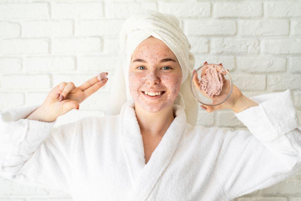 Woman in white bathrobe and towel applying natural face scrub, holding bowl, smiling