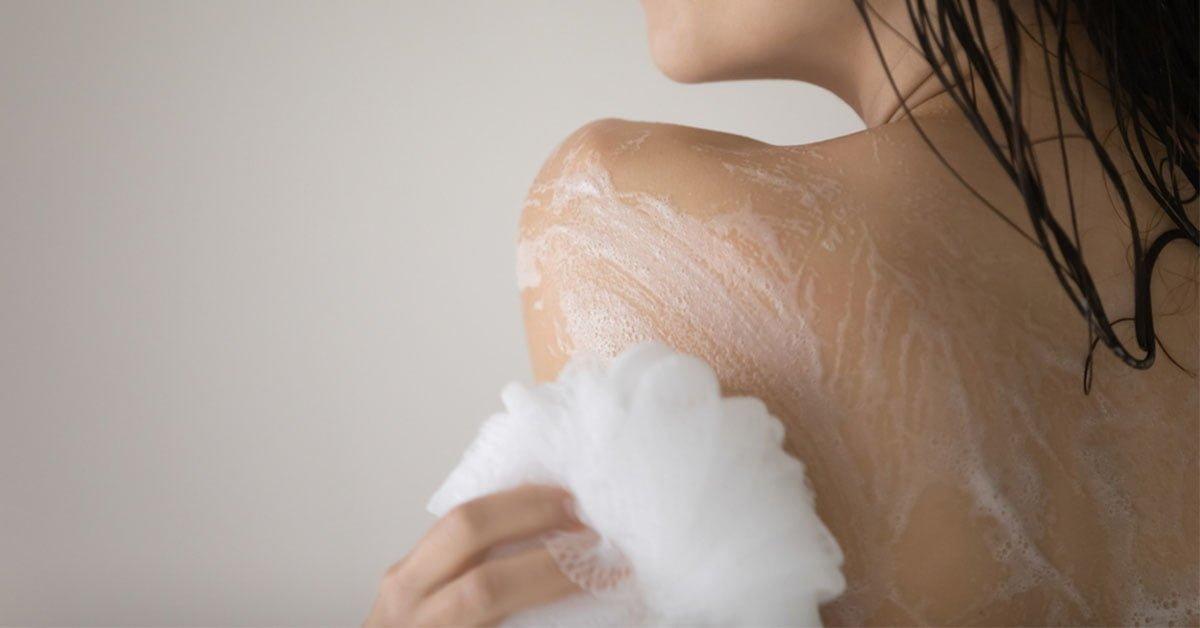 Woman using bath sponge to lather soap on shoulder, skincare and bathing routine