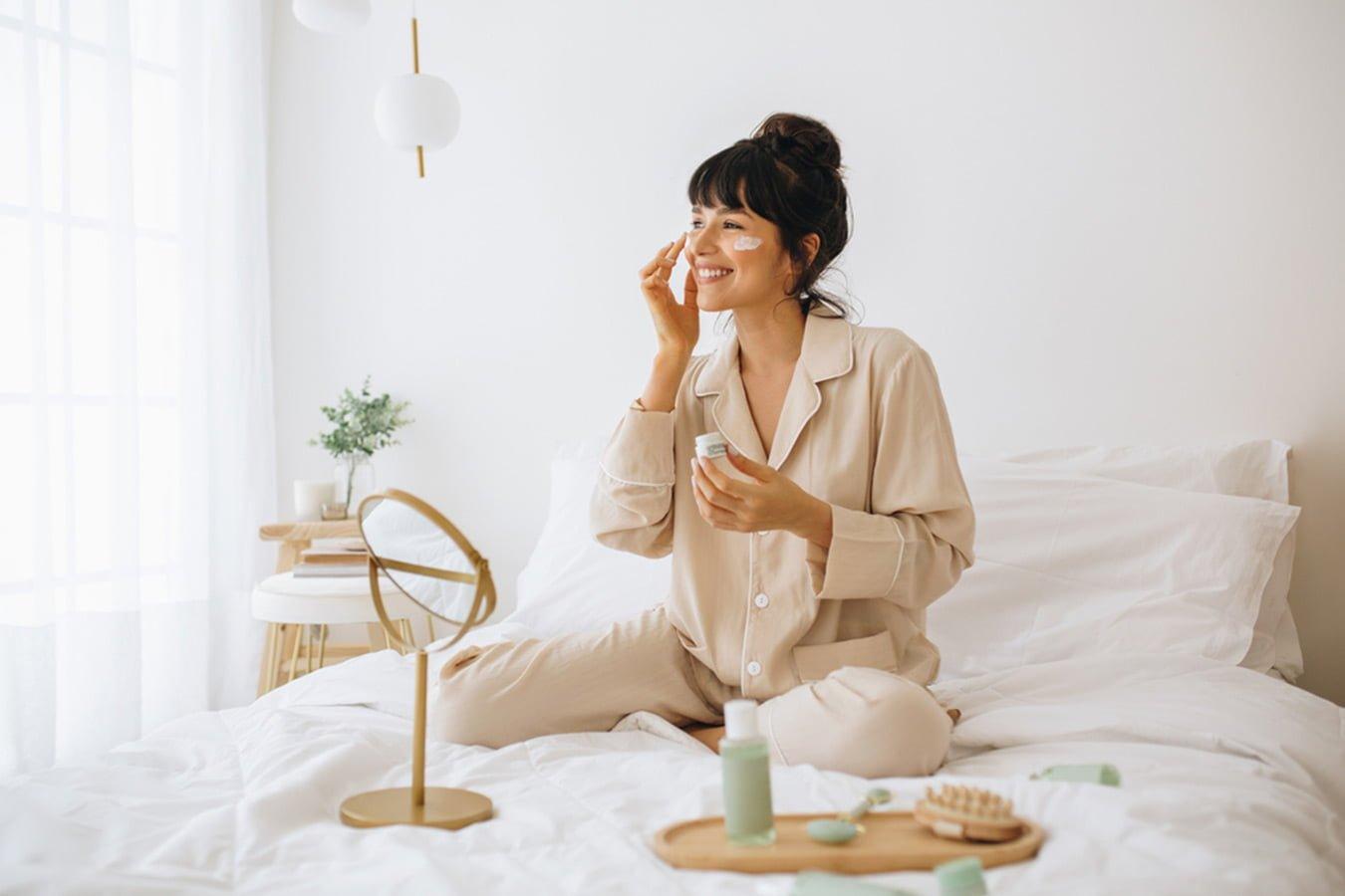 Woman in pajamas applying natural skincare cream while sitting on bed in bright bedroom