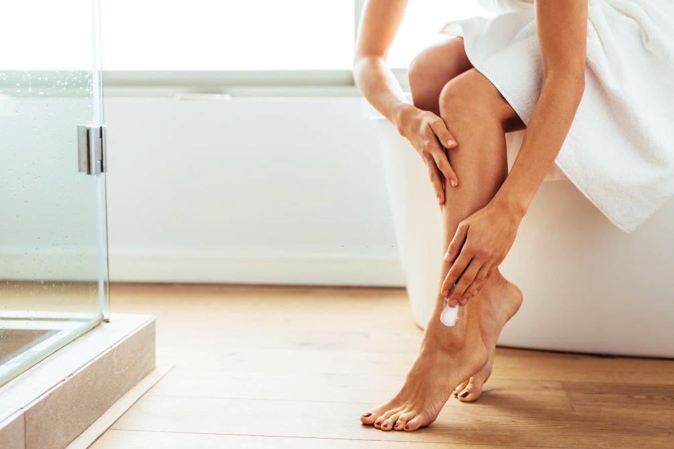 Woman applying herbal body lotion to legs after shower in bright bathroom