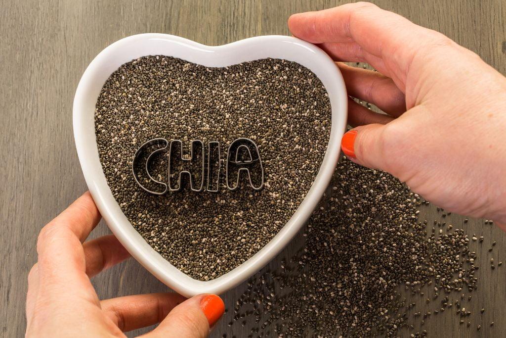 Heart-shaped bowl filled with chia seeds on a wooden table, held by hands with orange nails