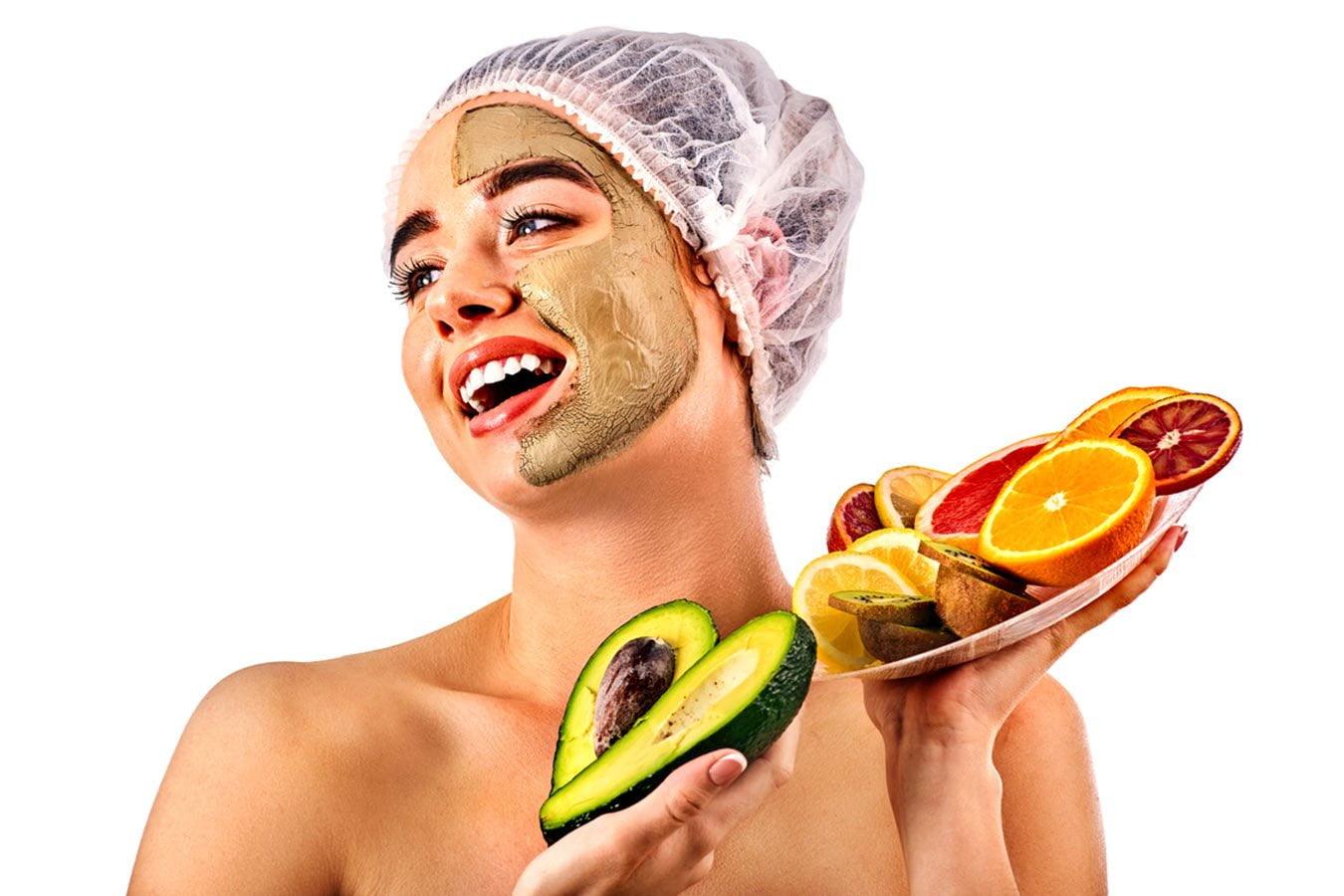 Woman with clay face mask, shower cap, holding avocado and plate of fresh citrus fruits