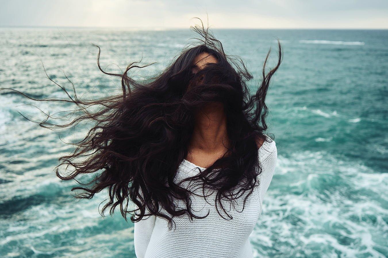 Woman with long, wavy hair in wind near ocean, natural hair care setting