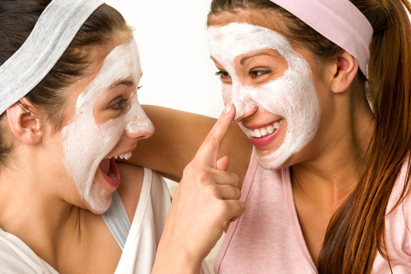 Two women with white face masks and headbands laughing together, enjoying skincare routine