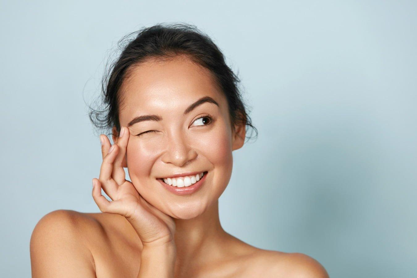Smiling woman with glowing skin winking, promoting natural skincare, on light blue background
