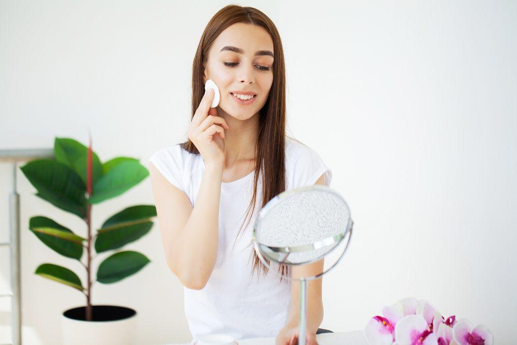 Woman applying skincare with cotton pad in front of mirror, bright indoor setting with plants