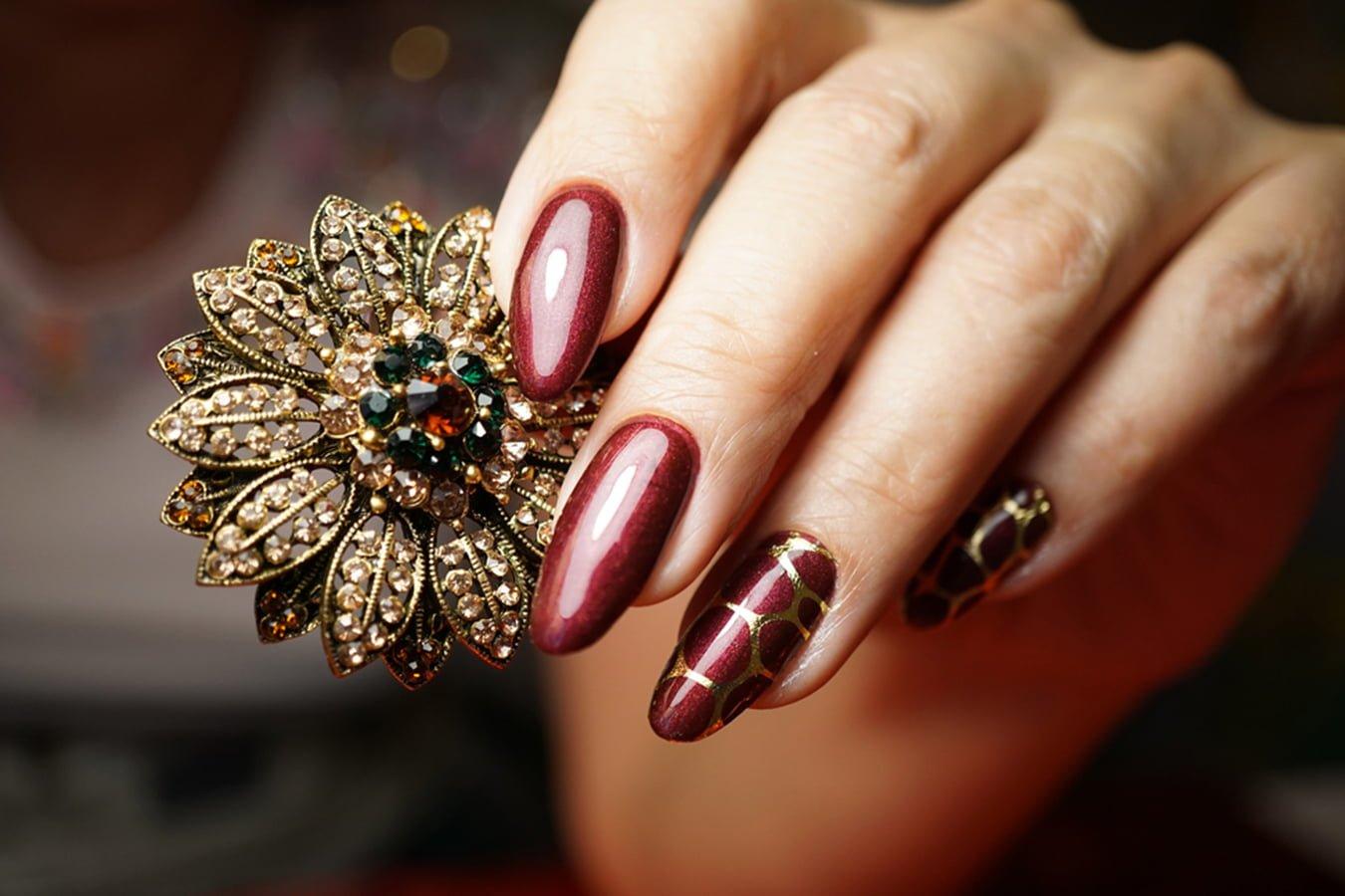 Hand with burgundy and gold nail art holding an ornate jeweled brooch
