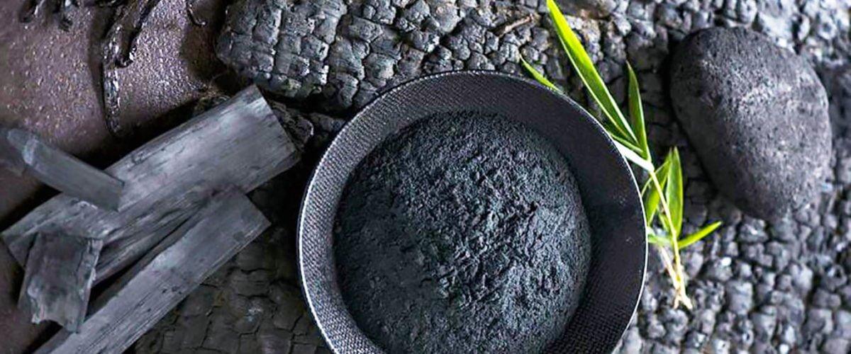 Activated charcoal powder in a bowl with charcoal pieces and green leaves on a textured surface