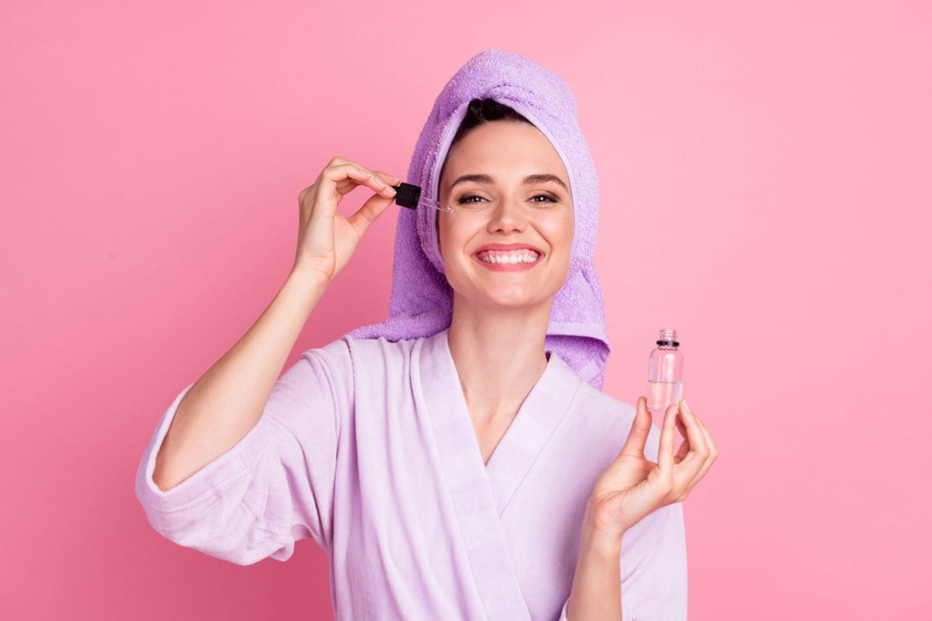 Smiling woman in a bathrobe and towel applying facial serum with a dropper on pink background