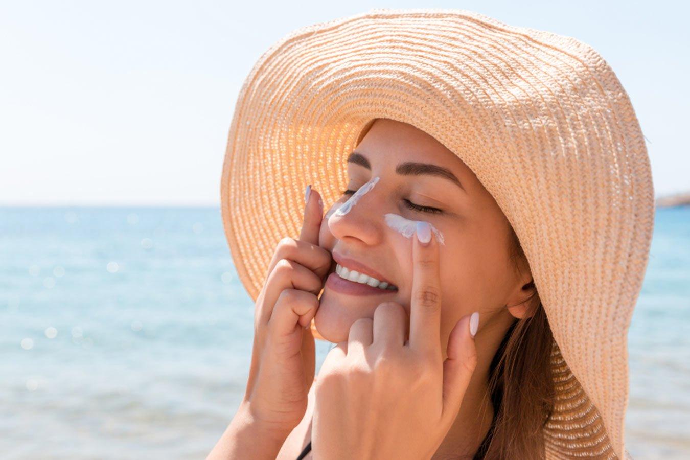 Woman applying sunscreen lotion on face at the beach, wearing a sun hat, summer skincare