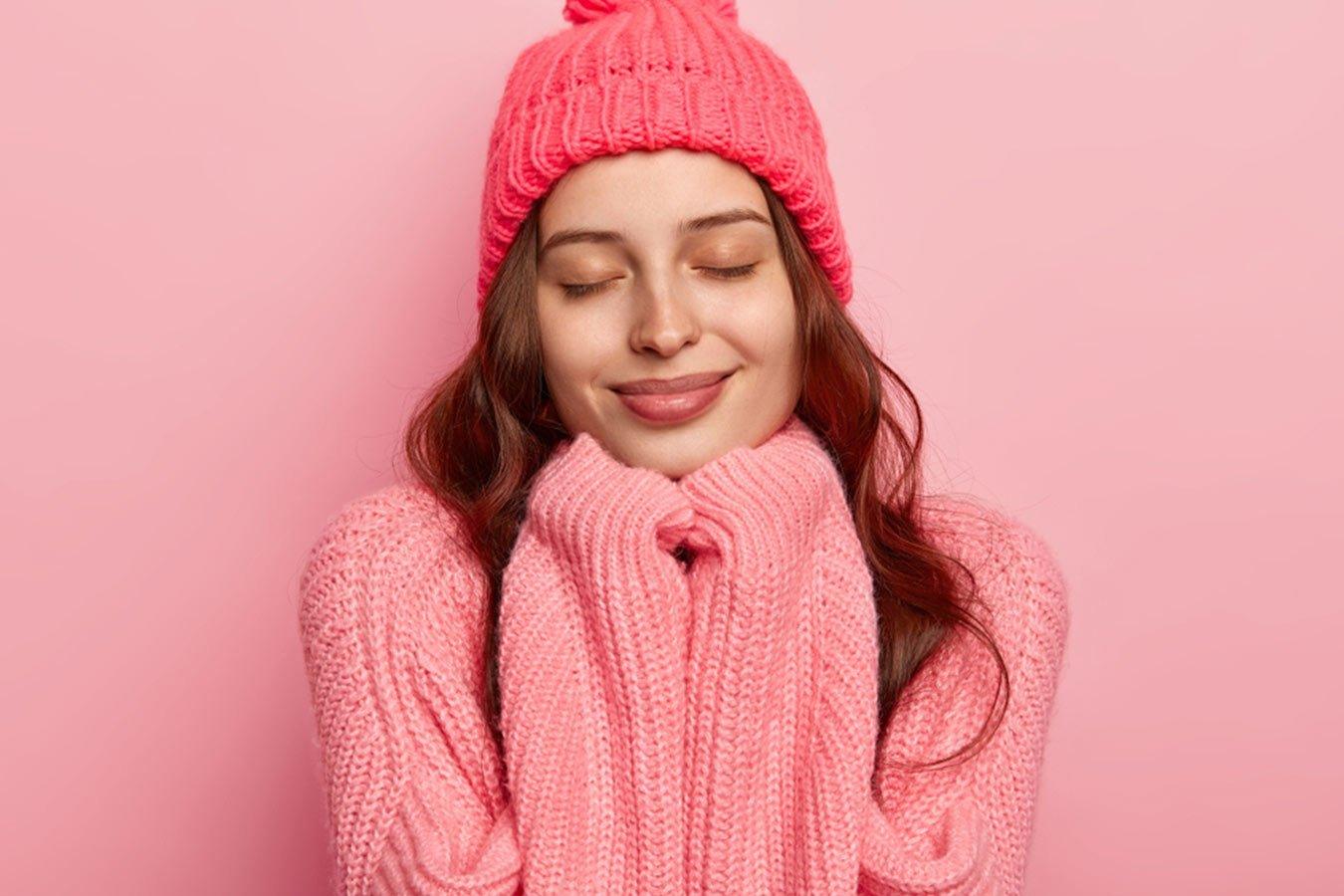 Woman in pink knitted sweater and beanie, cozy winter skincare, smiling on pink background