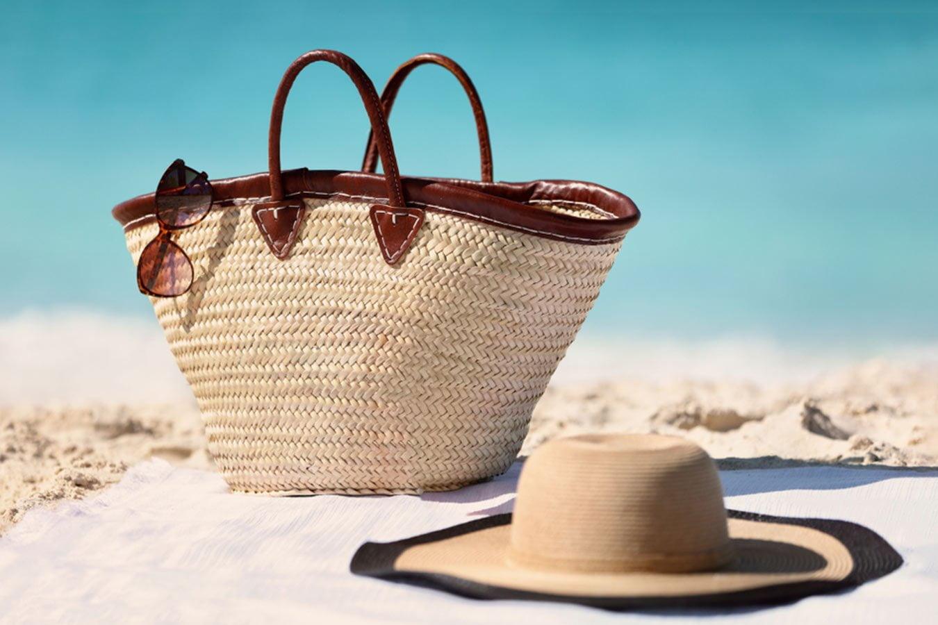 Straw beach bag with sunglasses and sun hat on sandy beach by clear blue sea