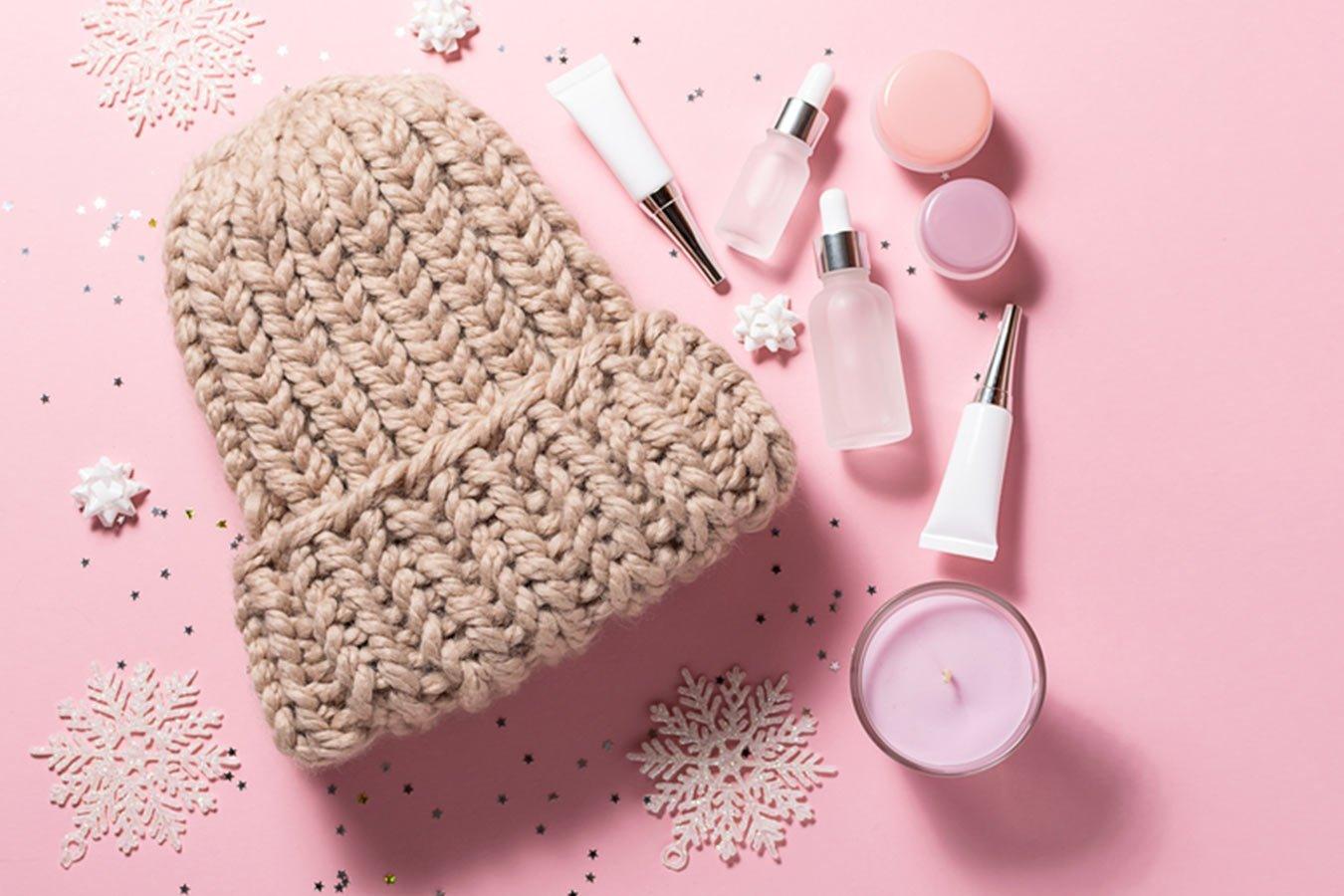 Winter skincare products, knitted wool hat, candle and snowflakes on pink background