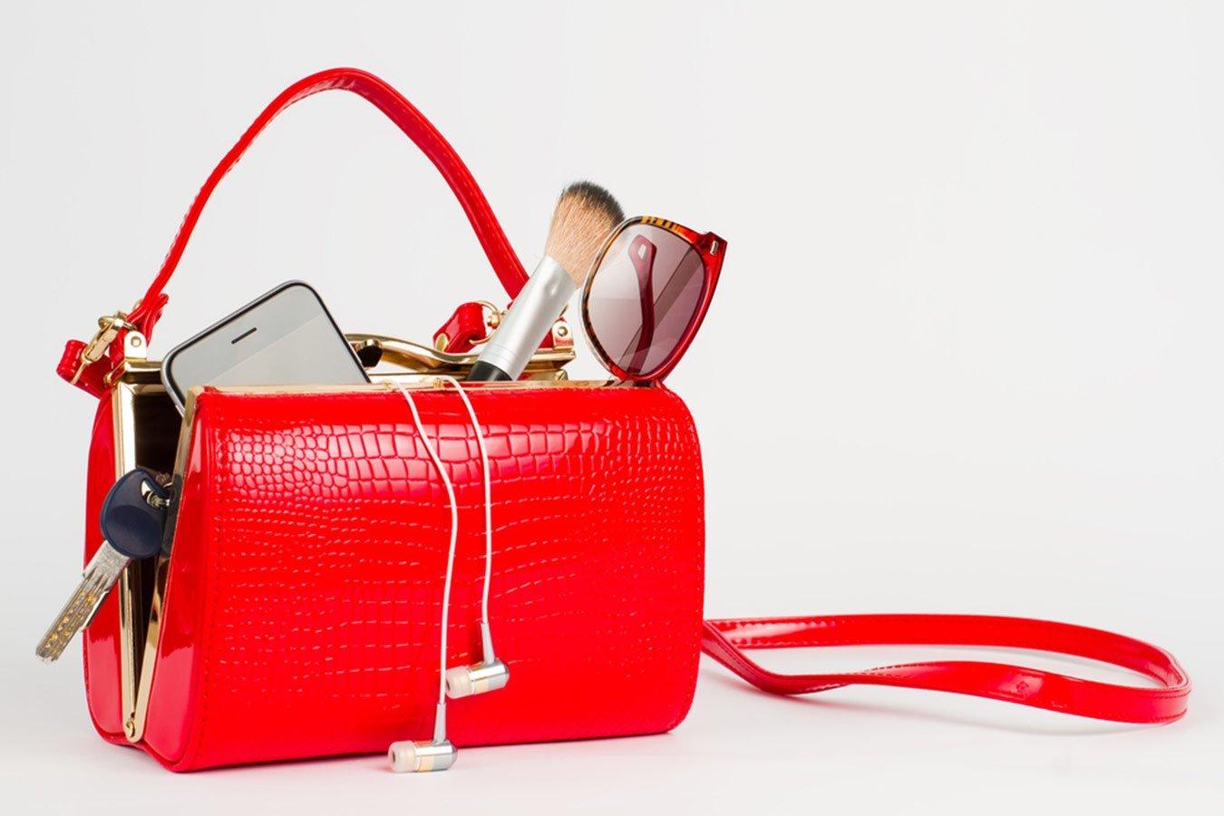 Red textured handbag with sunglasses, makeup brush, smartphone, earphones, and keys spilling out