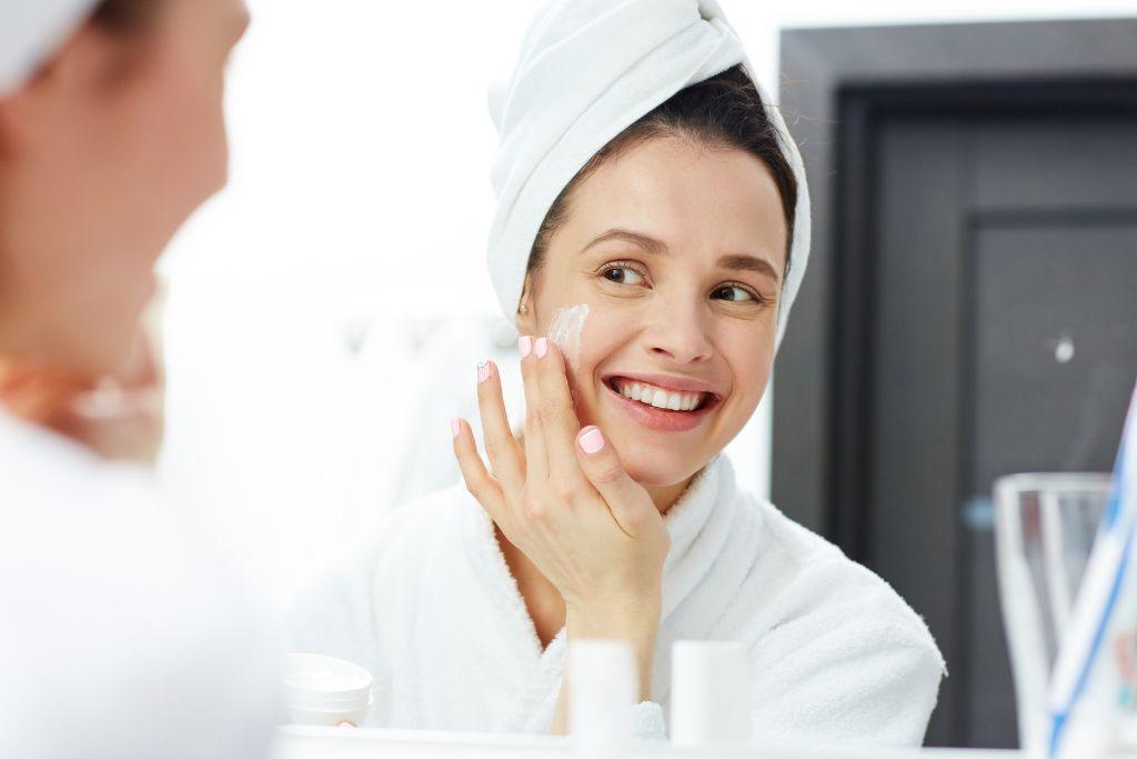 Woman applying cleansing milk to face in bathroom, wearing white robe and towel head wrap