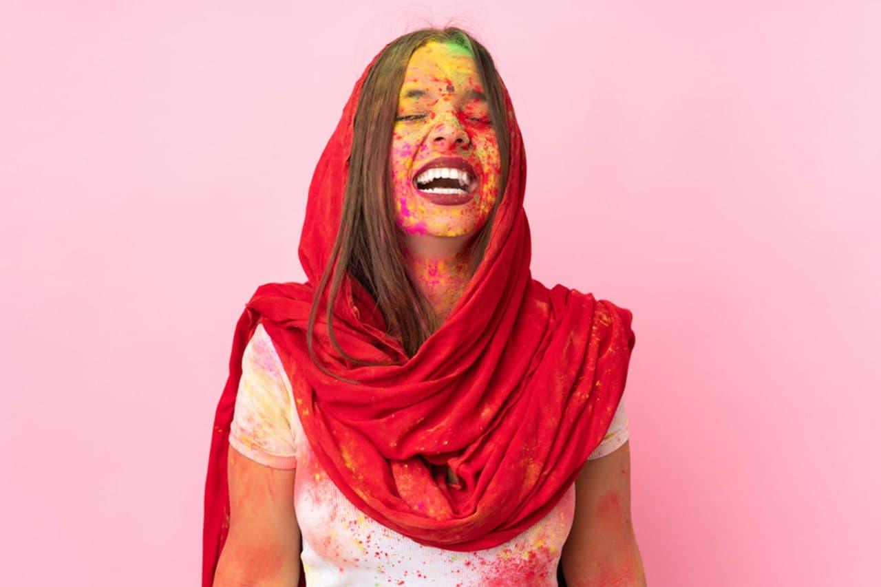 Smiling woman in red scarf celebrating Holi with colorful powder on face against pink background